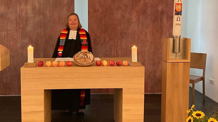Eine Pfarrerin im schwarzen Talar steht hinter einem mit Obst und Kerzen geschmückten Altar und blickt lächelnd in die Kamera.