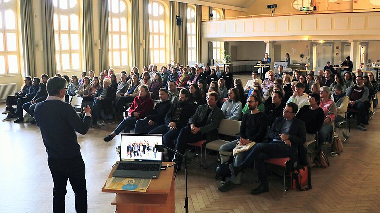 Ein Mann hält vor ca. 80 Zuhörerinnen und Zuhörern einen Vortrag.