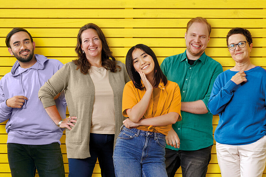 Eine Gruppe von Menschen, jung und alt, mit bunter Kleidung steht vor einer gelben Holzwand.