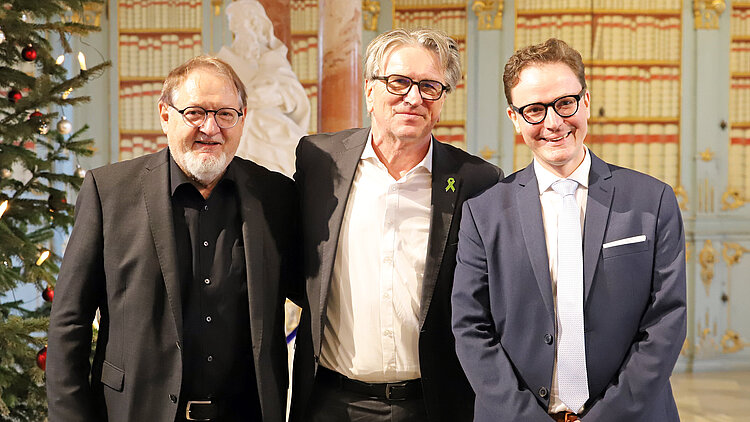 Drei Männer in schicken Anzügen und Hemd stehen gemeinsam in einer Bücherei vor einem geschmückten Weihnachtsbaum.
