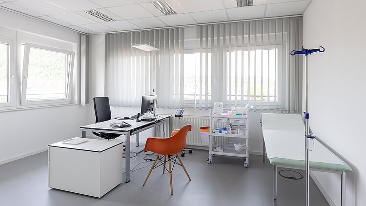 Ein Praxiszimmer mit Schreibtisch, orangenem Besucherstuhl und einer Behandlungsliege.